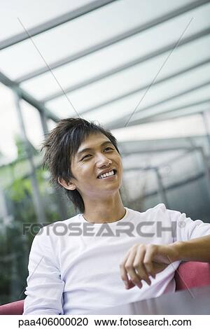 Young man sitting at table, looking away and smiling View Large Photo Image Stock Image - Young man sitting at table, looking away and smiling. Fotosearch