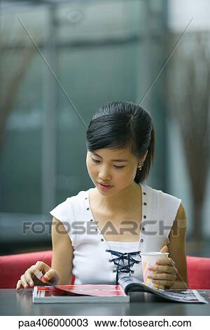 Young woman sitting at table, looking at magazine, holding drink View Large Photo Image Stock Image - Young woman sitting at table, looking at magazine, holding drink. Fotosearch