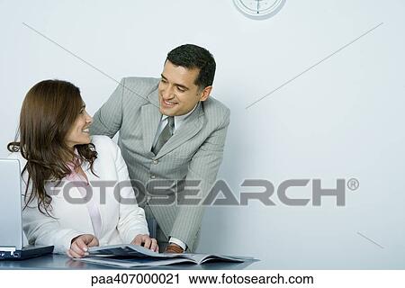 Businessman and businesswoman smiling at each other while woman sits at desk and man looks over her shoulder View Large Photo Image Stock Image - Businessman and businesswoman smiling at each other while woman sits at desk and man looks over her shoulder. Fotosearch