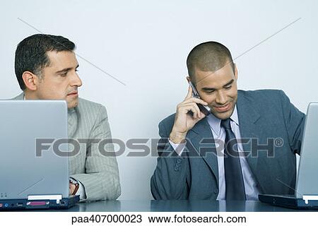 Two businessmen using laptops, one using cell phone while other watches him View Large Photo Image Stock Image - Two businessmen using laptops, one using cell phone while other watches him. Fotosearch