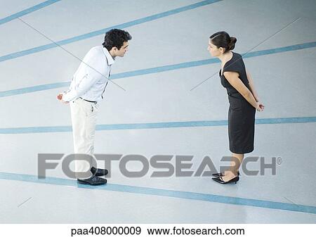 Stock Photo - Man and woman standing face to face, hands behind back, leaning toward each other, full length, high angle view. Fotosearch