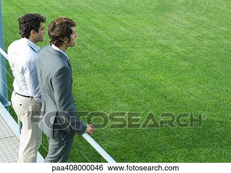 Two businessmen standing on walkway, looking into distance View Large Photo Image Stock Photograph - Two businessmen standing on walkway, looking into distance. Fotosearch