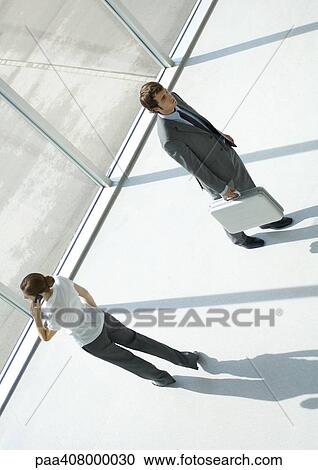 Stock Image - Woman using cell phone and man walking away with briefcase, full length, high angle view. Fotosearch