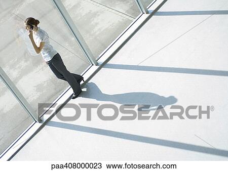 Stock Image - Young woman using cell phone by window, full length, high angle view. Fotosearch
