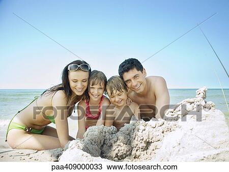 Stock Image - Family sitting on beach near pile of sand, portrait. Fotosearch