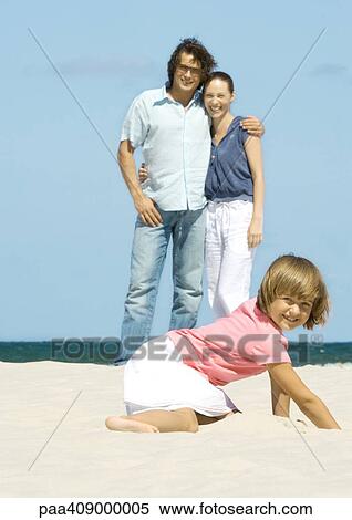 Girl digging in sand while parents stand in background View Large Photo Image Stock Photography - Girl digging in sand while parents stand in background. Fotosearch