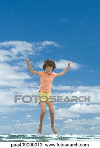Stock Image - Girl jumping, sky and ocean in background, full length. Fotosearch
