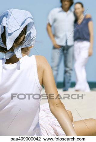 Stock Image - Girl sitting on beach, facing parents in background, rear view. Fotosearch