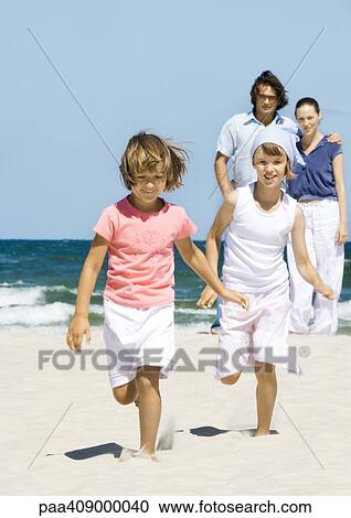 Stock Image - Girls running on beach while parents stand in background. Fotosearch
