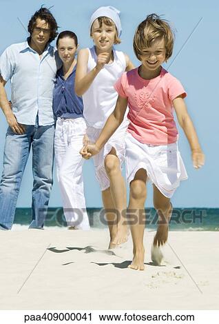 Stock Image - Girls running on beach while parents stand in background. Fotosearch