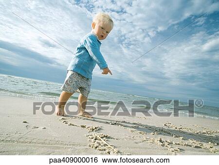 Stock Photograph - Toddler standing on beach, looking at camera and pointing at sand. Fotosearch