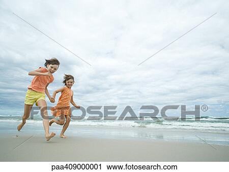 Stock Image - Two girls running hand in hand on beach, full length. Fotosearch