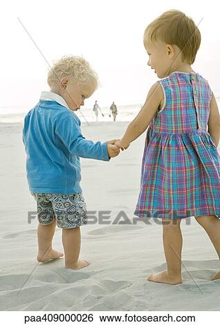 Stock Photograph - Two toddlers standing on beach, rear view. Fotosearch