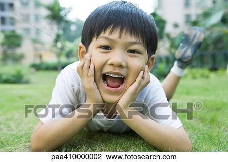 Stock Image - Boy lying on grass, holding head in hands, mouth wide open. Fotosearch