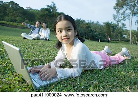 Girl lying in grass using laptop, parents in background View Large Photo Image Picture - Girl lying in grass using laptop, parents in background. Fotosearch