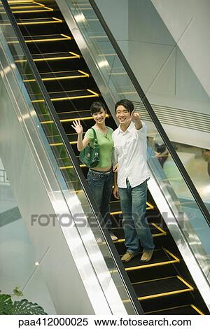 Stock Photography - Couple on escalator pointing at camera. Fotosearch
