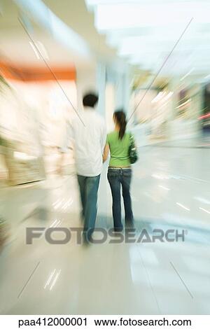 Stock Image - Couple walking through shopping mall, rear view, full length. Fotosearch