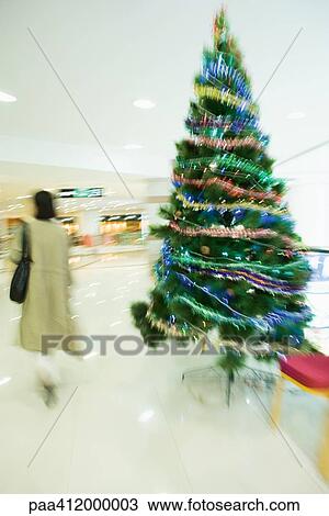Stock Image - Female shopper walking past Christmas tree in mall. Fotosearch