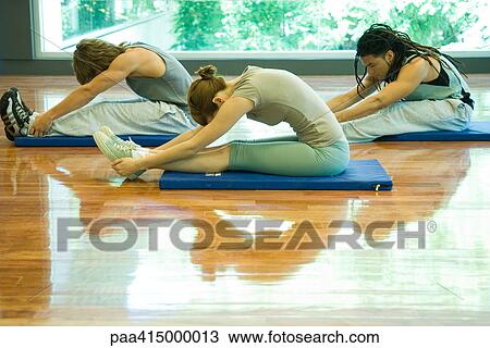 Stock Image - Exercise class doing seated forward bend, side view. Fotosearch