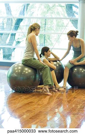 Stock Image - Young women sitting on fitness balls, chatting, in gym room. Fotosearch