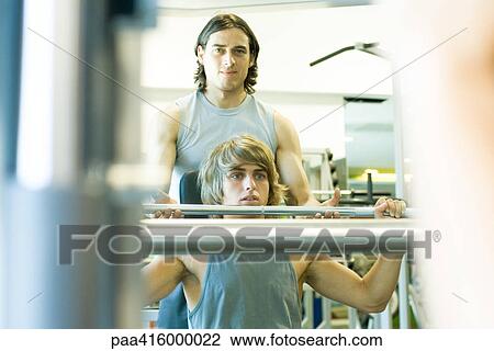 Stock Image - Man lifting barbell, second man spotting. Fotosearch