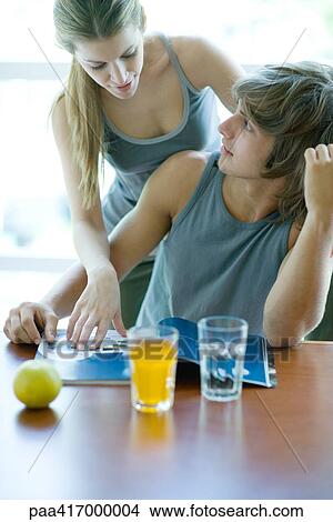 Picture - Young adults in exercise clothing having snack in health club cafeteria, looking at magazine. Fotosearch