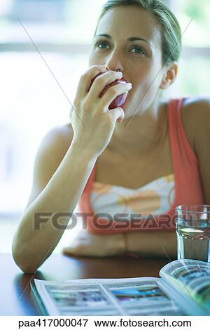Stock Photo - Young woman sitting with magazine and glass of water, eating apple. Fotosearch