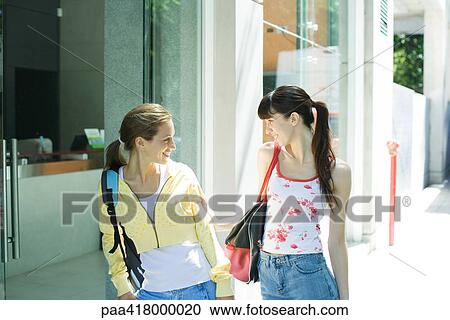 Stock Image - Two young women standing on sidewalk, looking at each other. Fotosearch