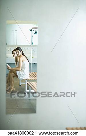 Stock Image - Two young women wrapped in towels, sitting on bench in locker rooms, reflected in mirror. Fotosearch