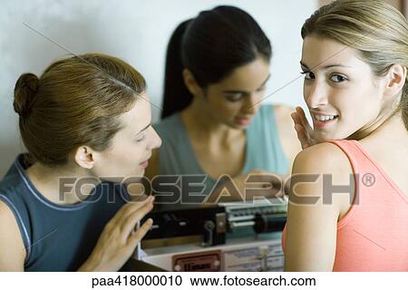 Stock Image - Woman standing on scale while friends look at weight. Fotosearch