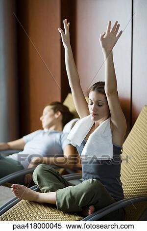 Stock Photography - Young women relaxing in lounge chairs, one sitting indian style with arms raised and eyes closed. Fotosearch