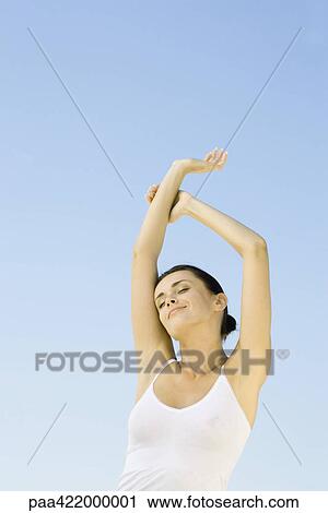 Stock Image - Woman stretching arms overhead, sky in background, low angle view. Fotosearch