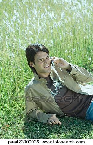 Stock Photography - Man lounging in field, using cell phone, eyes closed. Fotosearch