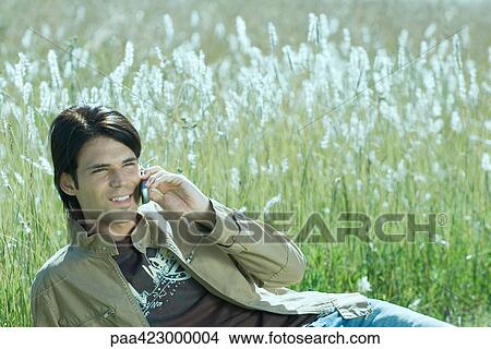 Young man reclining in field, talking on cell phone View Large Photo Image Picture - Young man reclining in field, talking on cell phone. Fotosearch