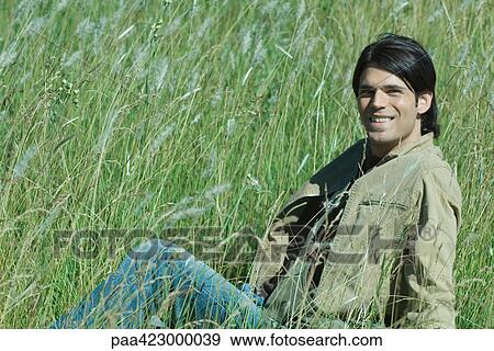 Young man sitting in long grass in field, smiling View Large Photo Image Stock Photo - Young man sitting in long grass in field, smiling. Fotosearch