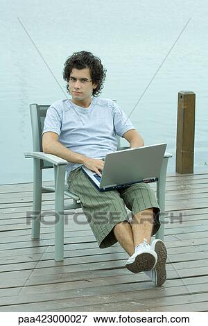 Young man sitting on dock using laptop, full length View Large Photo Image Stock Photo - Young man sitting on dock using laptop, full length. Fotosearch