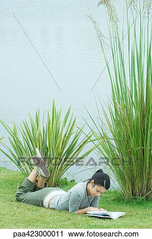 Stock Image - Young woman lying on grass near body of water, reading. Fotosearch