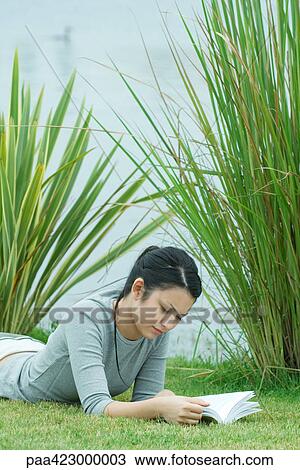 Stock Image - Young woman lying on grass, reading. Fotosearch