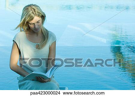 Picture - Young woman sitting by water, reading. Fotosearch