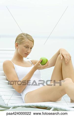 Stock Image - Teenage girl in swimsuit sitting on lounge chair, holding apple. Fotosearch