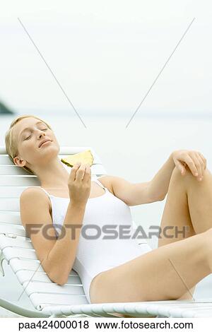 Stock Photo - Teenage girl in swimsuit sitting on lounge chair, holding slice of pineapple. Fotosearch