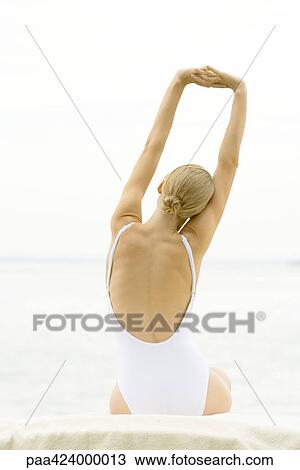 Stock Image - Teenage girl sitting stretching arms, wearing bathing suit, rear view. Fotosearch