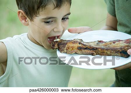 Stock Image - Boy eating grilled meat from plate. Fotosearch