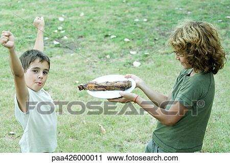 Stock Image - Boy handing friend plate of grilled meat. Fotosearch