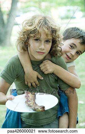 Stock Image - Boy holding grilled meat on plate, friend riding piggy back. Fotosearch