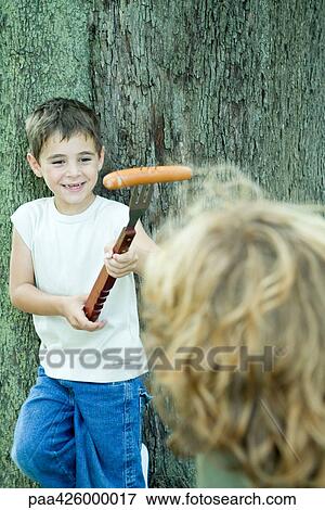 Stock Photo - Boy holding hot dog on end of large fork, showing friend . Fotosearch
