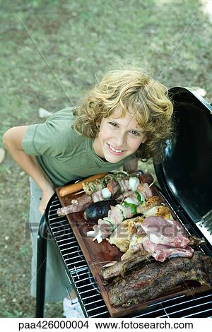 Picture - Boy leaning over grilling food, smiling at camera. Fotosearch