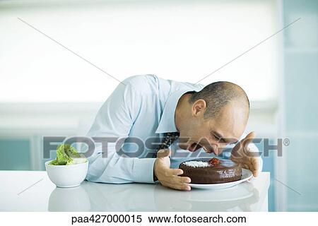 Man sitting at table with chocolate cake and bowl of lettuce, leaning toward cake with mouth wide open View Large Photo Image Stock Photography - Man sitting at table with chocolate cake and bowl of lettuce, leaning toward cake with mouth wide open. Fotosearch