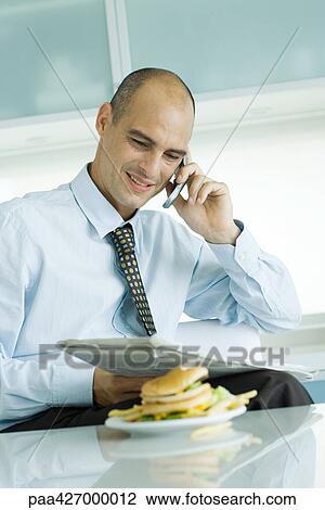 Man using phone and reading newspaper, hamburger on table in foreground View Large Photo Image Stock Image - Man using phone and reading newspaper, hamburger on table in foreground. Fotosearch