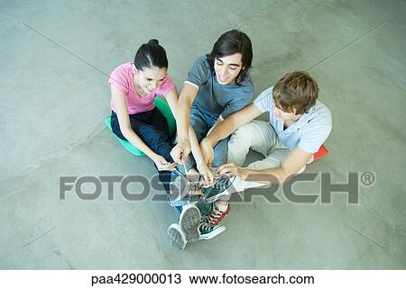 Stock Image - Teenage friends with legs joined at ankles, tying laces of canvas shoes. Fotosearch